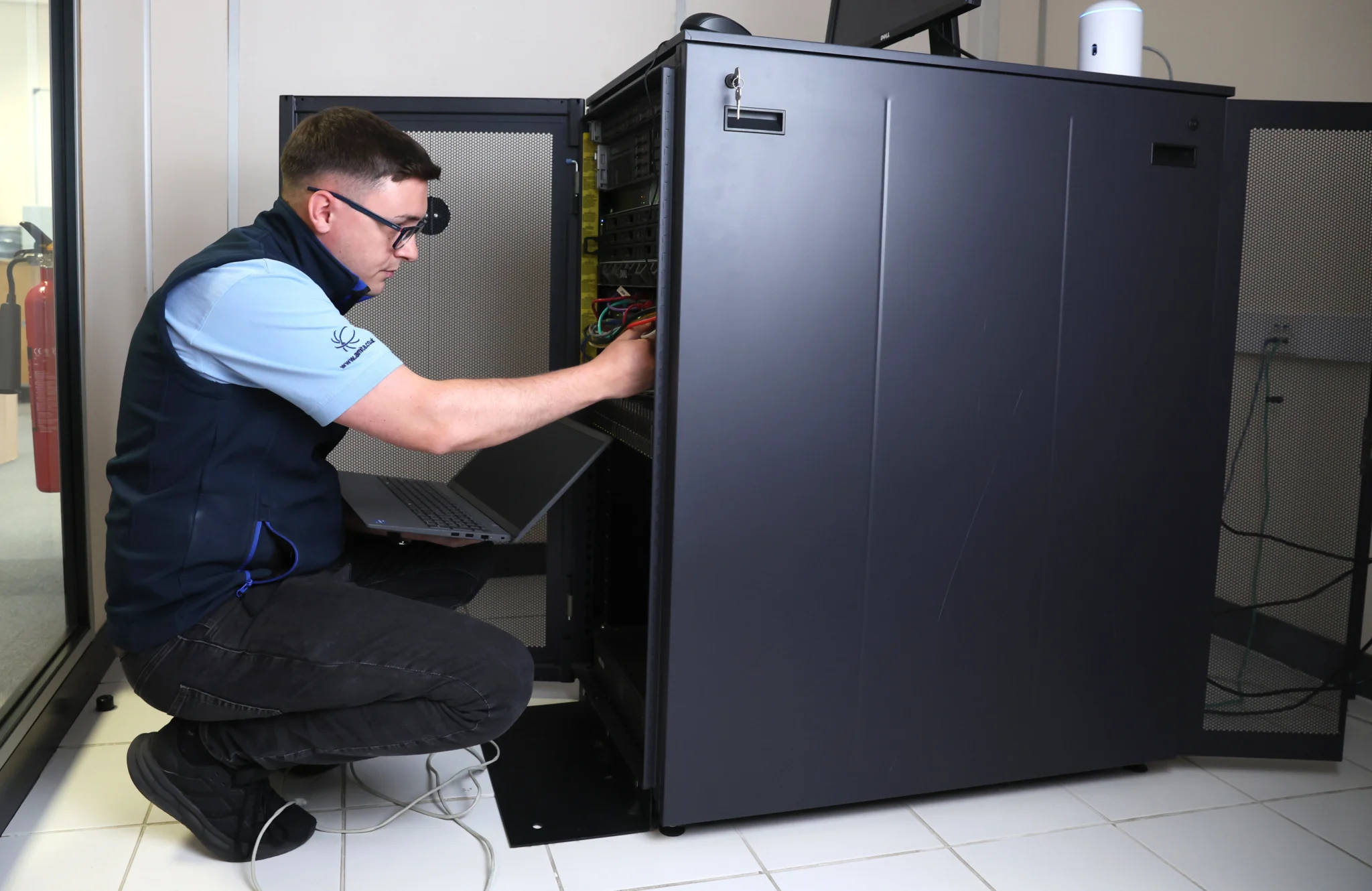A technician in a blue shirt and vest crouches beside a server rack, using a laptop and checking cables. The setting is a well-lit server room, conveying focus and professionalism.