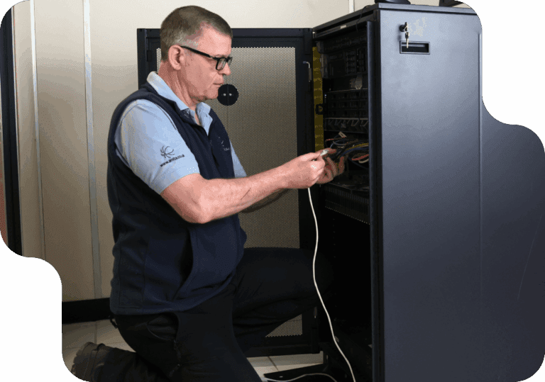 Man kneeling and working on a server rack, holding a cable. He's dressed in a blue uniform, focused, in a technical room, conveying professionalism.