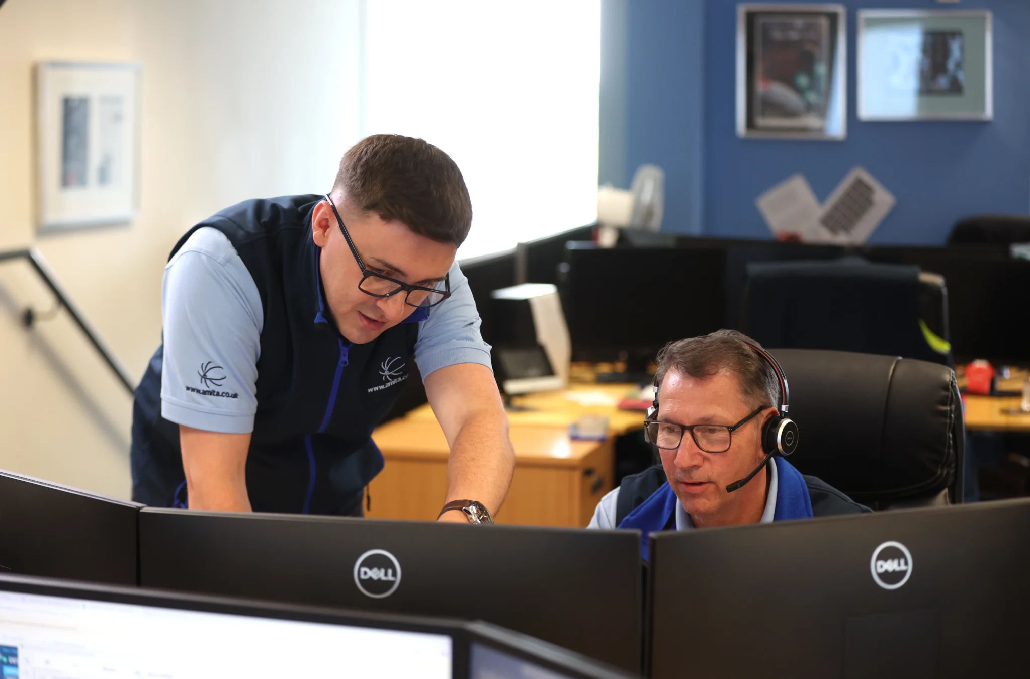 Two men in an office, one standing and pointing at a computer screen, the other seated with headphones, engaged in focused discussion.