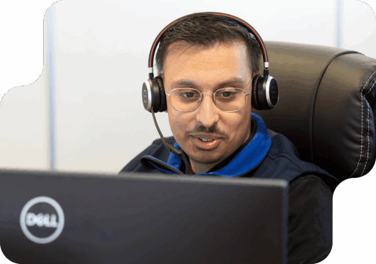 A person wearing glasses and a headset sits at a desk, focused on a computer screen with a Dell logo. The setting appears professional and engaged.