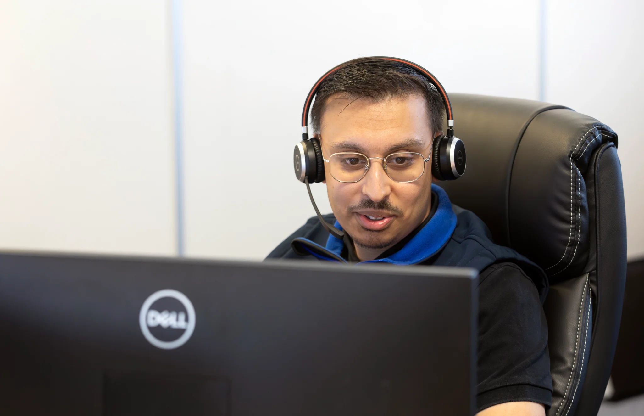 A person wearing glasses and a headset sits at a desk, focused on a computer screen with a Dell logo. The setting appears professional and engaged.