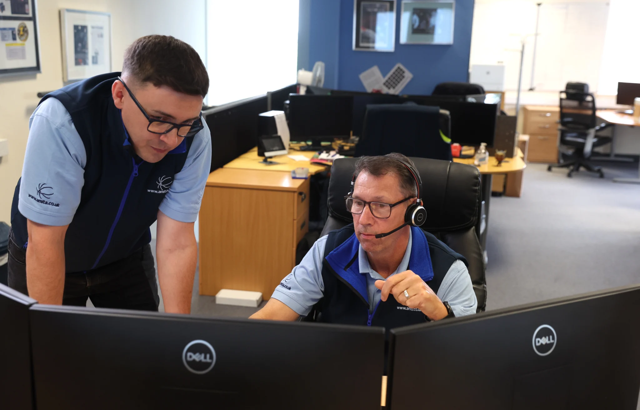 Two men in an office, both in blue company-branded shirts and vests, focus on computer monitors. One, providing IT support with a headset, is seated while the other stands beside him.