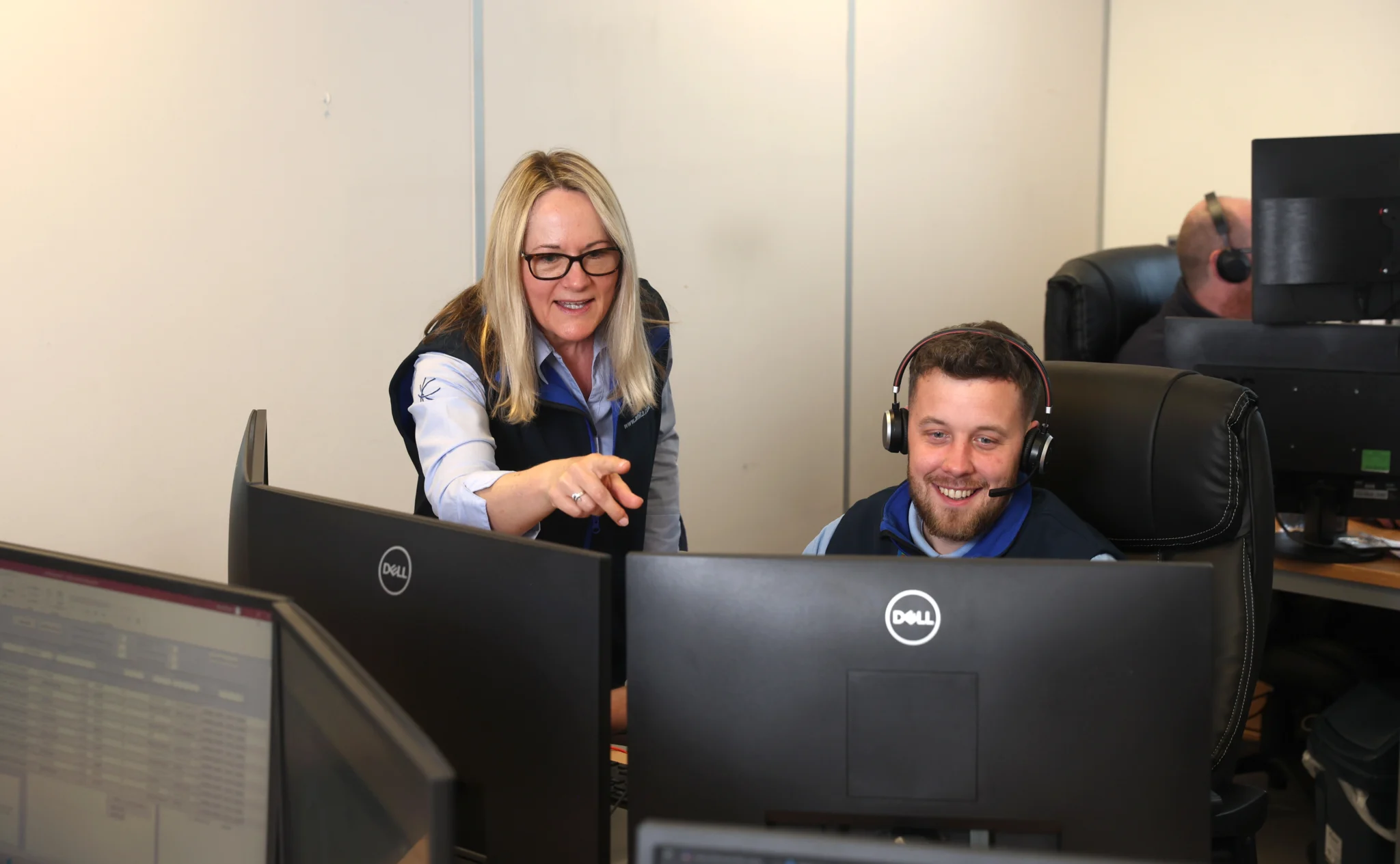 A woman stands and points at a computer monitor, providing IT support to a man wearing a headset who sits and looks at the screen in an office with multiple monitors.